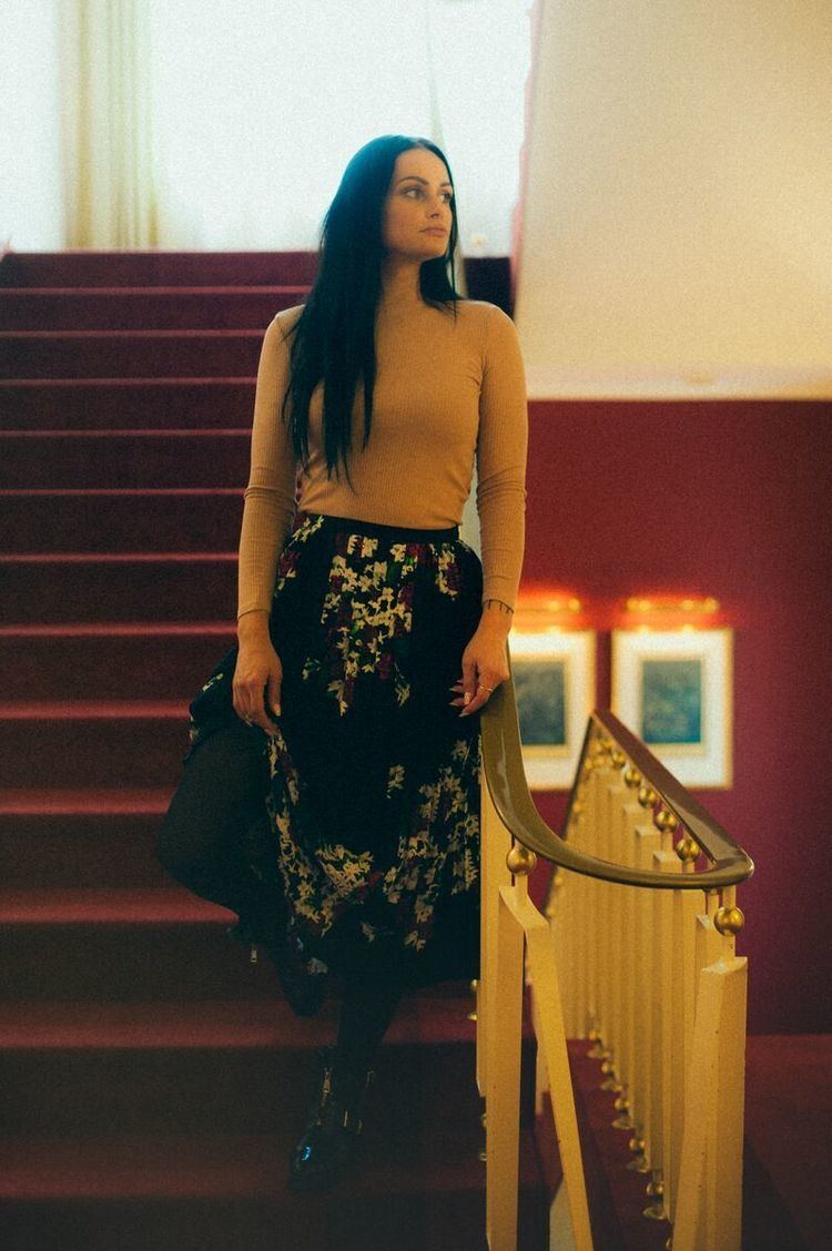 Yazmeen Baker looking afar while at the stairs and wearing a beige knitted long sleeve blouse and black skirt with floral design