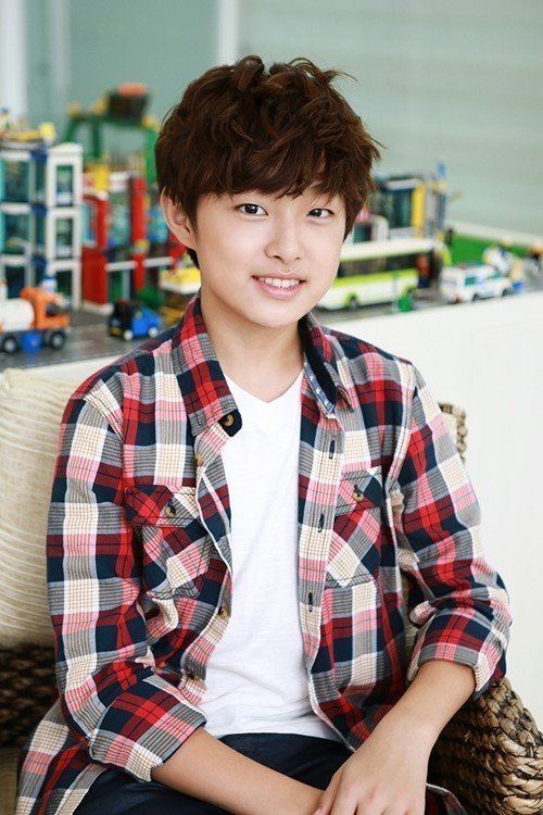 Yoon Chan-young smiling, sitting, and holding his hands with cars and other toys in the background, is a South Korean actor. He has a brown messy hairstyle wearing a white V-neck shirt and a checkered long sleeve.