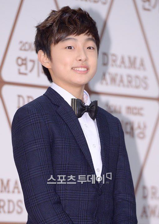 Yoon Chan-young smiling and standing on a carpet, a South Korean actor attending the 2014 MBC Drama Awards at MBC in December 2014, in Seoul, South Korea, with a brown messy hairstyle wearing a bowtie on a white collared long sleeve and a dark blue plaid coat.