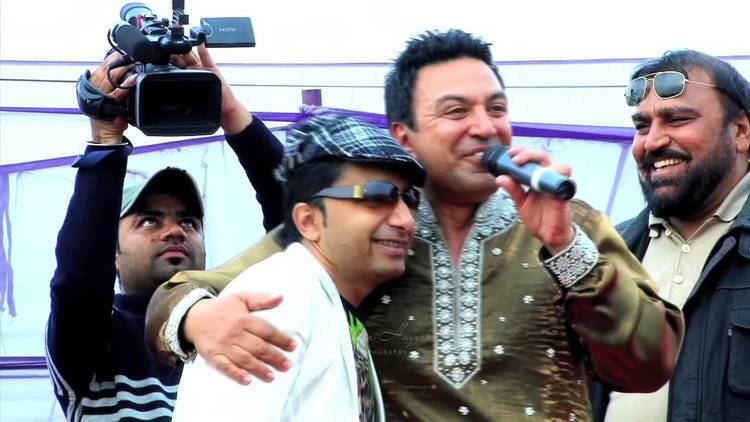 Manmohan Waris holding a microphone and Yudhvir Manak smiling while wearing checkered hat, shades and white coat