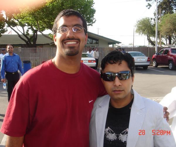 Yudhvir Manak wearing shades, white coat and black shirt and a man beside him smiling and wearing eyeglasses and red shirt