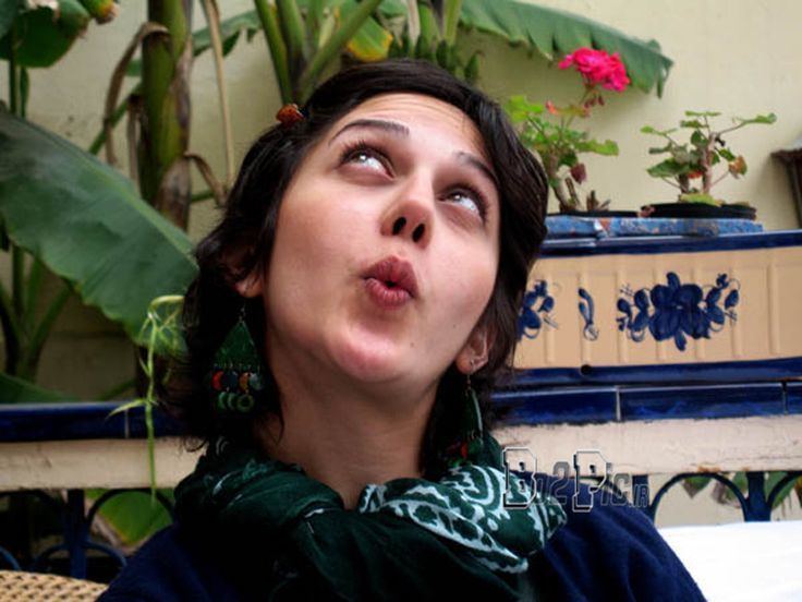 Zahra Amir Ebrahimi  whistling while looking above with her black hair and plants in her background, she is wearing earrings, a green scarf on her neck, and a dark blue blouse