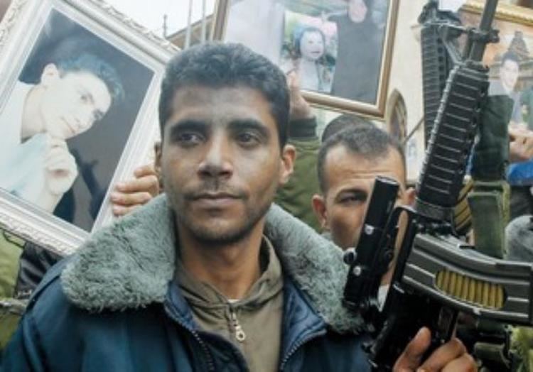 Zakaria Zubeidi is surrounded by people with guns while he is wearing a gray and blue fur jacket