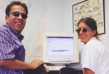 Ziad Jarrah smiling while wearing black shades and violet checkered polo and the woman beside him wearing a white blouse and shades