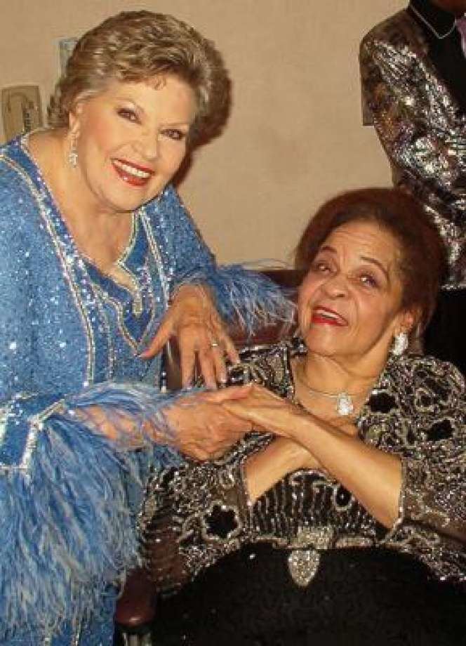 Zola Taylor smiling and wearing a black glitter dress with a lady wearing a blue dress.