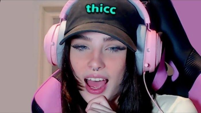 Zookdook sitting on a black and pink gaming chair with her mouth open and she is wearing pink headphone, a black cap, and a circular barbell